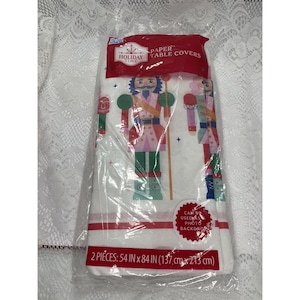 May include: A package of two Holiday Time paper table covers measuring 54 inches by 84 inches. The white covers feature a repeating pattern of colorful nutcracker figures in red, green, and blue, with small star accents and red border stripes.