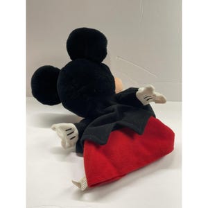 May include: A plush Mickey Mouse toy, featuring a black head and ears, a tan face, and white gloves. The toy is wearing a black coat and red shorts. The toy is lying on a white surface.