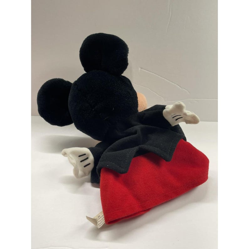 May include: A plush Mickey Mouse toy, featuring a black head and ears, a tan face, and white gloves. The toy is wearing a black coat and red shorts. The toy is lying on a white surface.
