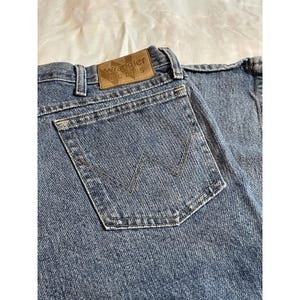 May include: Close-up of blue denim jeans with a back pocket featuring a stitched 'W' design. The jeans have a brown leather patch with the 'Wrangler BLUES' logo. The denim has a faded appearance, suggesting wear.