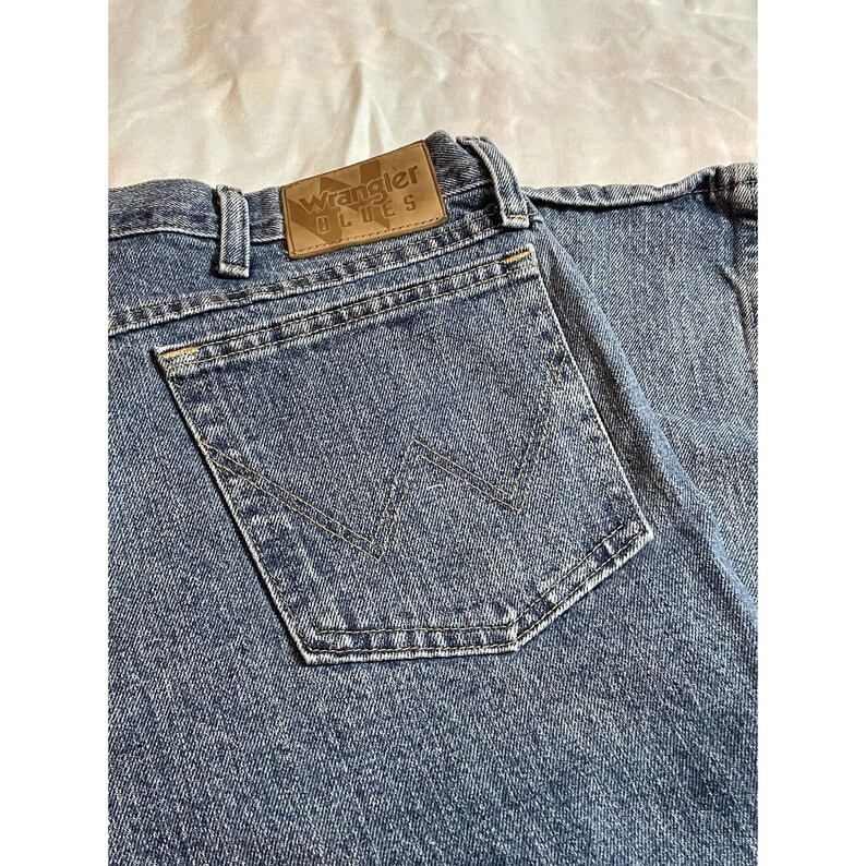 May include: Close-up of blue denim jeans with a back pocket featuring a stitched 'W' design. The jeans have a brown leather patch with the 'Wrangler BLUES' logo. The denim has a faded appearance, suggesting wear.