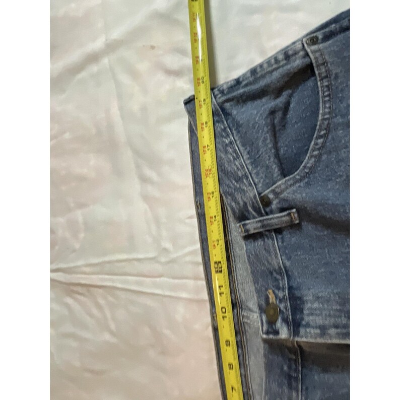 May include: A light blue denim jacket with a button closure and a small pocket. The jacket is being measured with a yellow tape measure, showing a length of approximately 11 inches.