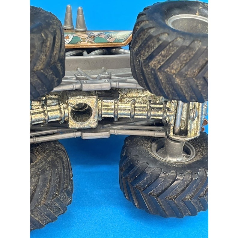 May include: A close-up view of a toy monster truck, showcasing its large, black, textured tires and detailed chassis. The truck has a silver and gold color scheme with some decorative elements on the top.