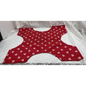 May include: A red fabric piece with a repeating pattern of small white hearts and tiny white dots. The textile has a distinct cutout shape with a curved neckline and four extended corners, featuring a flat, symmetrical design throughout.