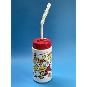 May include: A white plastic drinks bottle with a red lid and a white, flexible straw. The bottle is decorated with yellow, red, and black geometric shapes and the words "Yeah!" and "Kool-Aid".