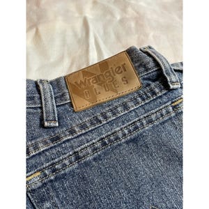 May include: Close-up of blue denim jeans with a brown leather patch. The patch is embossed with the words "Wrangler BLUES". The jeans have visible stitching and a classic design.