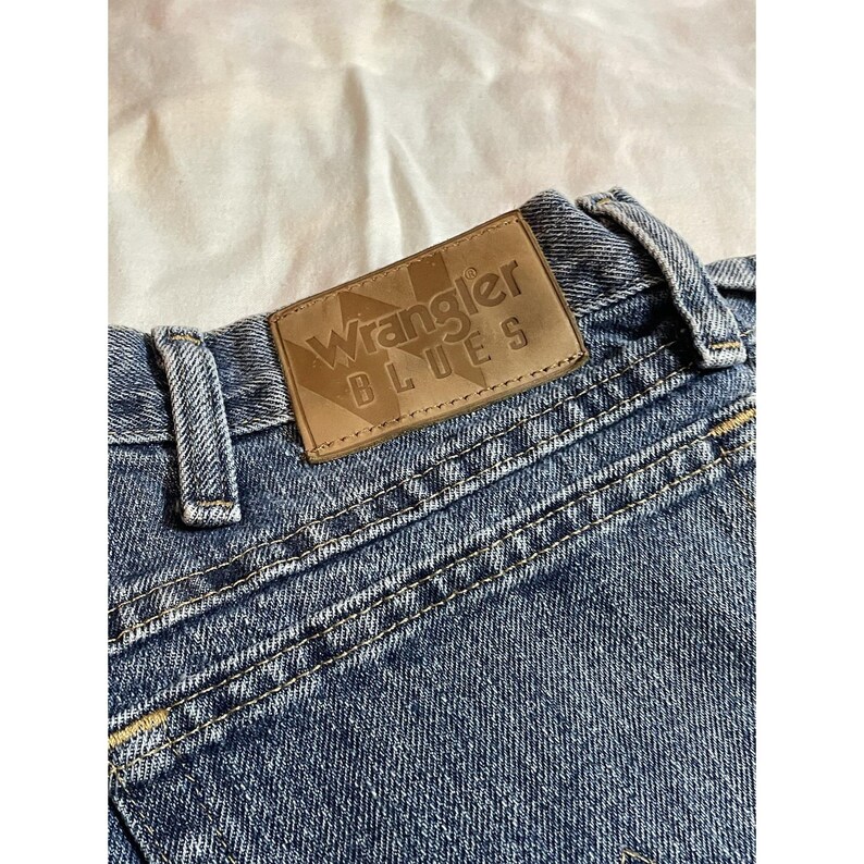 May include: Close-up of blue denim jeans with a brown leather patch. The patch is embossed with the words "Wrangler BLUES". The jeans have visible stitching and a classic design.