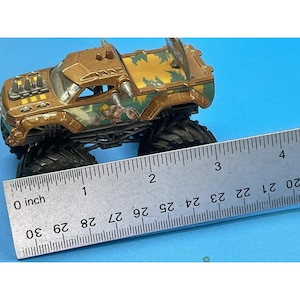 May include: A brown and green toy monster truck with large black tires. The truck has a He-Man graphic on the side and measures approximately 3 inches long.