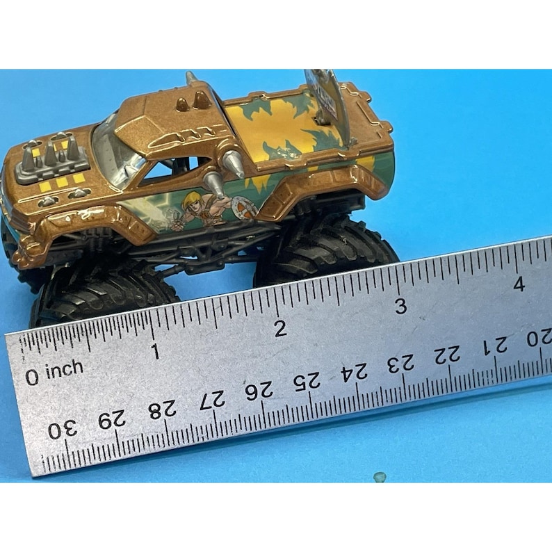 May include: A brown and green toy monster truck with large black tires. The truck has a He-Man graphic on the side and measures approximately 3 inches long.