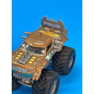 May include: A brown and gold toy monster truck with large black tires. The truck features the text "MASTERS OF THE UNIVERSE" on the top and has a battle axe design. The toy has a blue and green trim.