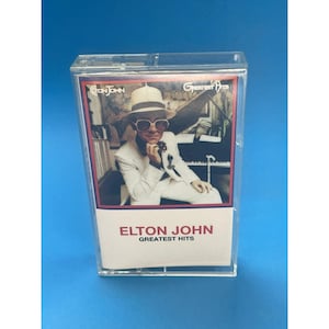 Può includere: Una musicassetta vintage di "Greatest Hits" di Elton John. La copertina dell'album presenta una foto di Elton John con occhiali da sole e un abito bianco. La cassetta è racchiusa in un astuccio di plastica trasparente.