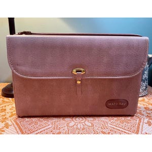 Könnte beinhalten: Eine rechteckige Kosmetiktasche in Mauve mit einem goldenen Verschluss und dem geprägten Schriftzug "MARY KAY" auf der Vorderseite.