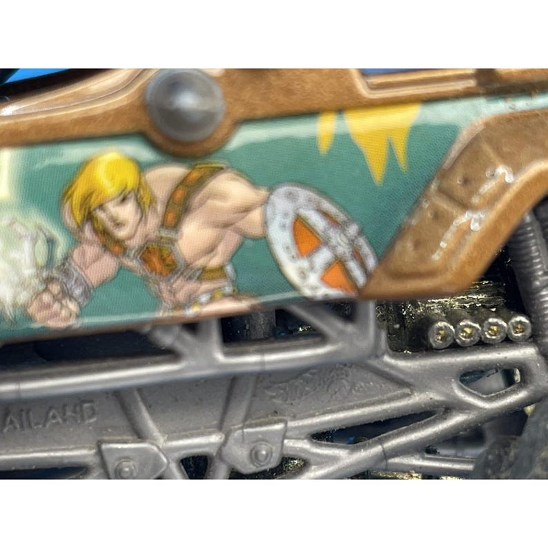 May include: Close-up of a toy truck featuring a cartoon image of He-Man. The truck has a silver chassis, brown and green accents, and a detailed illustration of the character on the side. The image is from the Hot Wheels brand.