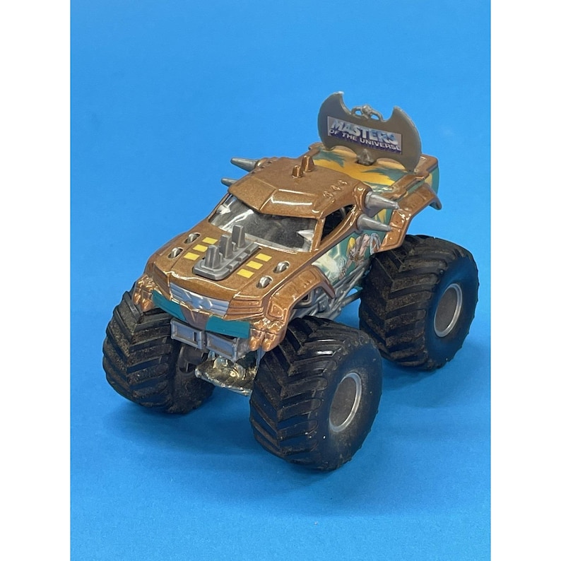 May include: A brown and silver toy monster truck with large black tires. The truck has the words "Masters of the Universe" on the top. The truck has a detailed design with various accents and a blue background.