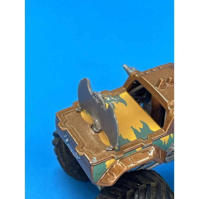 May include: A brown and yellow monster truck toy with a large, silver blade attached to the front. The truck has black tires and green and yellow accents. The background is a solid blue.