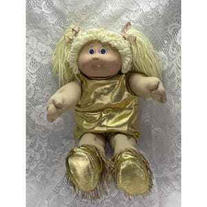 May include: A doll with light skin, blue eyes, and blonde yarn hair styled in two pigtails with small pink bows. The doll wears a metallic gold sleeveless top and matching gold shoes with fringe detailing around the ankles and a single tooth visible.