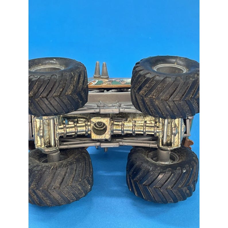 May include: A toy monster truck with large black tires and a detailed chassis. The truck is silver and brown with visible suspension components. The tires have deep treads for off-road use. The background is a solid blue.