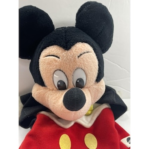 May include: A plush Mickey Mouse doll with black ears, a tan face, and large eyes. The doll wears a red outfit with yellow buttons and a white collar. The doll is a classic Disney character.