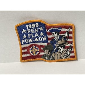 Pode incluir: Patch bordado com um desenho de bandeira com o texto "1990 PEN FLA POW-WOW" e ilustrações de figuras históricas com rifles. O patch tem um campo azul com estrelas brancas e um fundo listrado vermelho e branco, emoldurado por uma borda dourada.