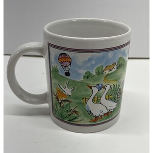 Puede incluir: Taza de cerámica blanca con una escena pastoral envolvente. El diseño presenta un globo aerostático, gansos, una casa y una valla. La taza tiene un asa blanca y un borde lila claro.