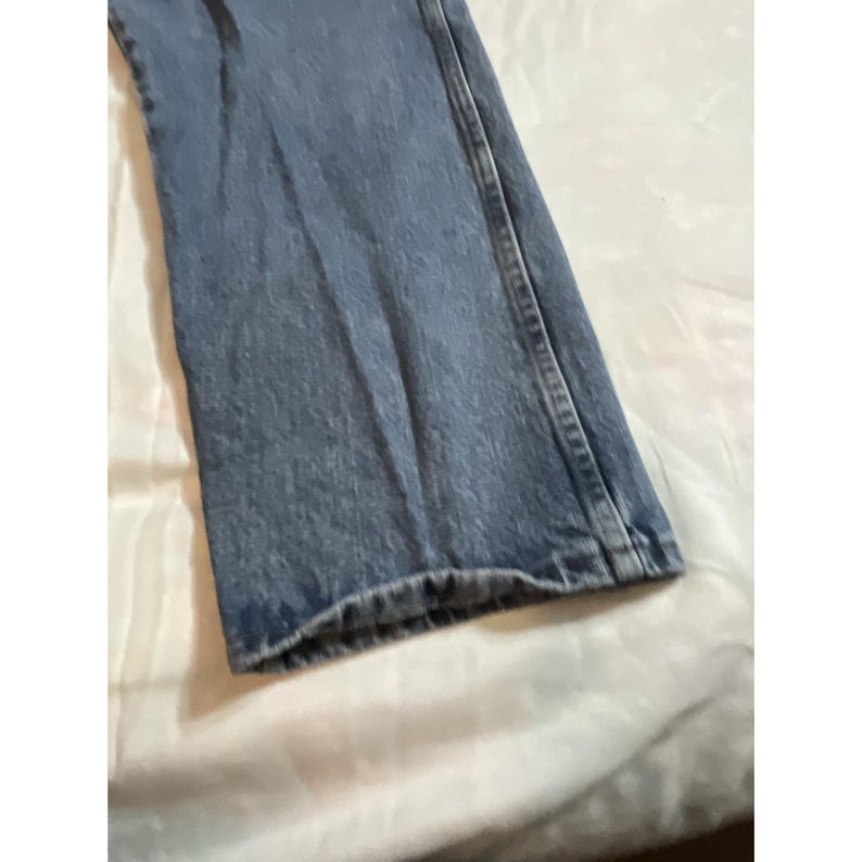 May include: Close-up of a section of blue denim jeans. The jeans are a light wash and appear to be slightly worn. The hem is visible, and the fabric has a textured appearance. The jeans are folded.
