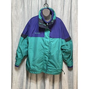 Könnte beinhalten: Eine Vintage-Jacke von Columbia Sports Company in Teal und Lila. Die Jacke hat einen hohen Kragen, einen Reißverschluss und das Columbia-Logo auf der Brust. Die Ärmel haben dunkelblaue Akzente. Die Jacke hängt an einem schwarzen Kleiderbügel.