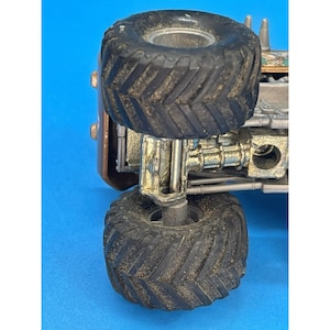 May include: Close-up of a toy monster truck with large, textured black tires. The truck has a bronze-colored body and metallic silver suspension components. The tires are covered in dirt, suggesting off-road play.