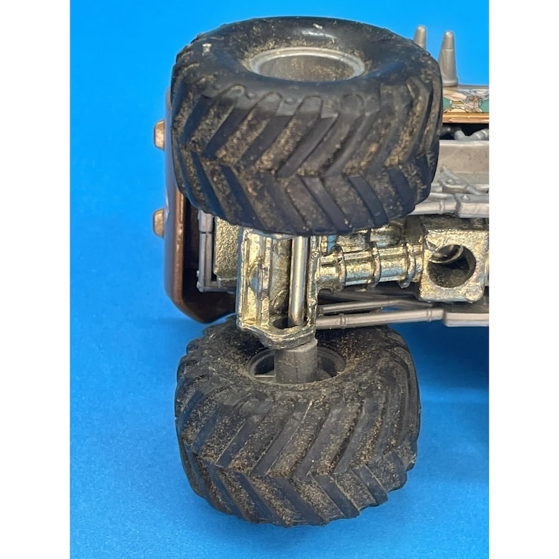 May include: Close-up of a toy monster truck with large, textured black tires. The truck has a bronze-colored body and metallic silver suspension components. The tires are covered in dirt, suggesting off-road play.