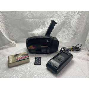 Puede incluir: Una videocámara Panasonic negra con visor extendido, un casete VHS-C Maxell TC-30 HGX-Gold, un pequeño mando a distancia negro y un cargador de batería negro con cable de alimentación, todo presentado junto como un conjunto.