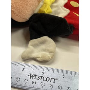 May include: A plush toy featuring a portion of a Mickey Mouse character. The toy has a tan face, black shorts, white gloves, and a red and yellow outfit. A ruler is placed next to the toy for measurement.