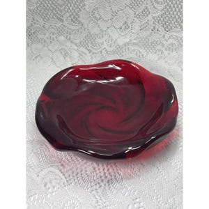Può includere: Un piatto in vetro rosso intenso a forma di rosa in fiore. La ciotola presenta un design traslucido e vorticoso con bordi curvi a forma di petalo che creano una forma circolare. Il vetro ha una superficie liscia e lucida con sfumature.