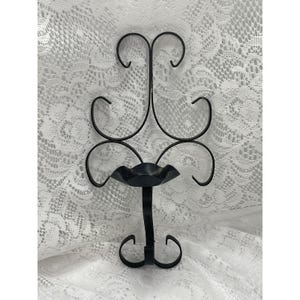 May include: A black metal wall sconce featuring a central candle holder plate with a ruffled edge. The frame is constructed from curved, scroll-like metal rods that extend upwards and downwards, creating a symmetrical, openwork design.