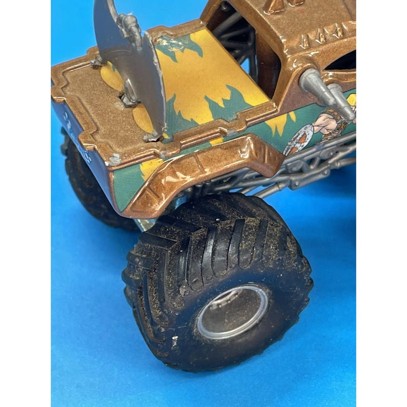 May include: A brown and gold toy monster truck with large black tires. The truck features a detailed design with green and yellow accents, and a cartoon graphic on the side. The truck has a metal blade on top.