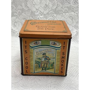 May include: A square metal tobacco tin with a tan lid labelled Continental Cubes Ready for the Pipe. The front features a graphic of a person in a cape and hat with the text Continental Cubes, flanked by the vertical phrases Its Cool and It Smokes.