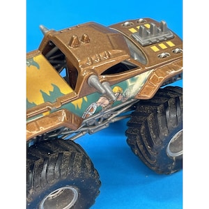 May include: A brown and gold monster truck toy with large black tires. The truck features a detailed design with spikes, a yellow and green graphic, and a cartoon character illustration on the side.
