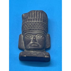 May include: A dark gray stone sculpture of a human head and shoulders. The figure wears a tall, cylindrical headdress featuring a textured, grid-like pattern on the lower band and vertical lines on the upper section. The face has almond-shaped eyes.
