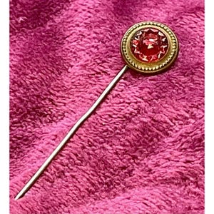 May include: A vintage hat pin with a round, gold-toned setting. The setting holds a faceted, red gemstone. The pin is long and slender, with a silver-colored finish. The background is a textured, pink fabric.