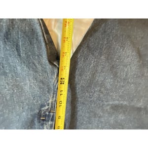 May include: Close-up of a blue denim vest with a button closure. A yellow measuring tape shows a length of approximately 10 inches. The vest has a classic design and appears to be well-worn.