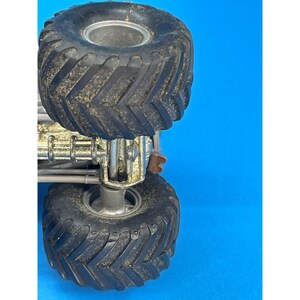 May include: Close-up of a toy monster truck with large, black, textured tires. The truck has a silver chassis and a blue background. The tires are covered in dirt, suggesting outdoor play.