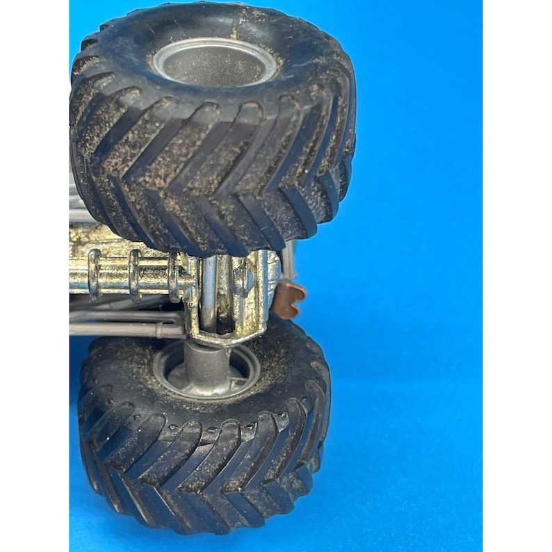 May include: Close-up of a toy monster truck with large, black, textured tires. The truck has a silver chassis and a blue background. The tires are covered in dirt, suggesting outdoor play.
