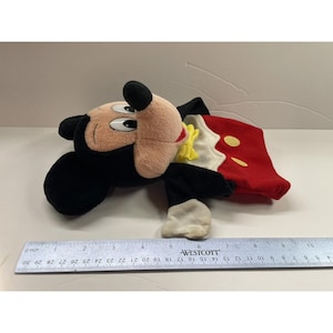 May include: A plush Mickey Mouse toy, featuring a black head and ears, a tan face, and a red and white outfit with yellow accents. The toy is lying on a white surface. A ruler is next to the toy.