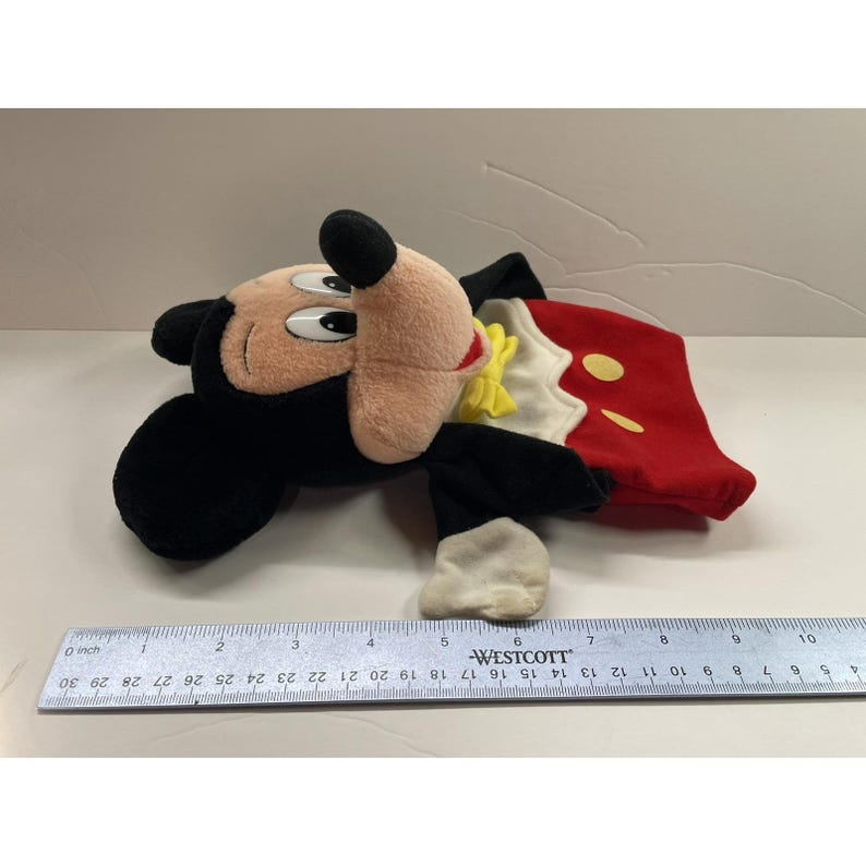 May include: A plush Mickey Mouse toy, featuring a black head and ears, a tan face, and a red and white outfit with yellow accents. The toy is lying on a white surface. A ruler is next to the toy.