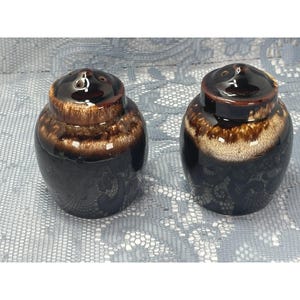 May include: Two ceramic salt and pepper shakers with a rounded shape. Each shaker features a dark brown base that transitions into a lighter, mottled tan glaze around the upper shoulder and a dark brown cap with small dispensing holes on top.