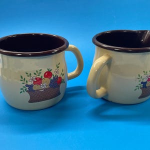 Op de afbeelding: Twee crèmekleurige keramische kannen met bruine randen en handvatten. Elke kan heeft een fruitmandontwerp met rode appels, paarse druiven en witte bloemen. Eén kan heeft een tuit. De kannen staan op een blauw oppervlak.