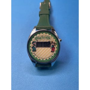 May include: Vintage Cabbage Patch Kids digital watch with a green leather strap. The watch face features the Cabbage Patch Kids logo and illustrations of two characters. The time displayed is 1:02. The watch has a silver-colored metal case.