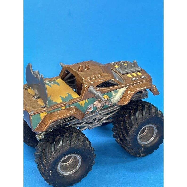 May include: A brown and green toy monster truck with large black tires. The truck features a detailed design with a character illustration on the side and a blue background.