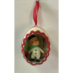 May include: A vintage Christmas ornament. A snowman figure is inside a cream-coloured egg-shaped shell with a red scalloped trim and a red ribbon. The snowman wears a black hat, green scarf, and red mittens. A festive decoration.