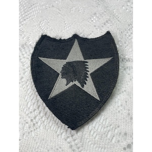 May include: A shield-shaped embroidered patch featuring a black background with a light gray five-pointed star in the center. Inside the star is a black silhouette profile of a person wearing a traditional feathered headdress, facing left.