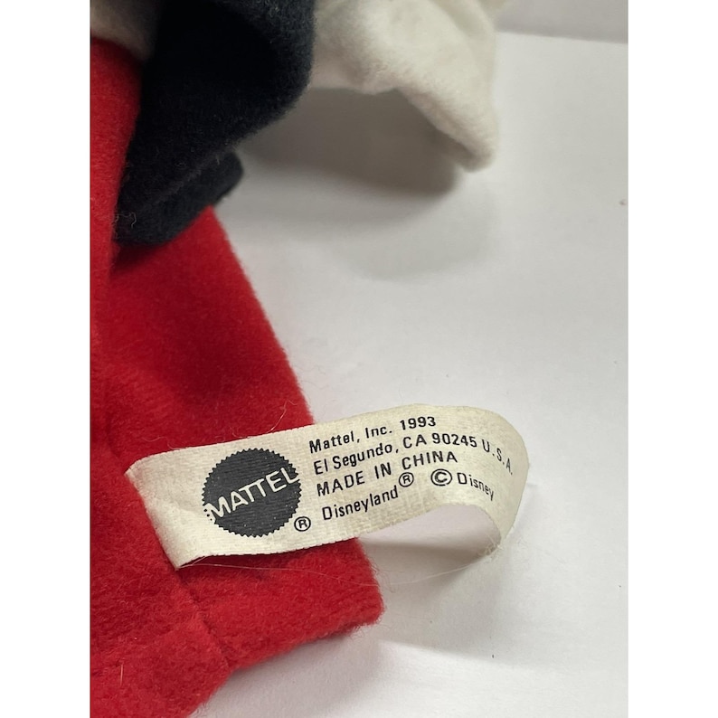 May include: A close-up of a vintage plush toy, featuring red, black, and white fabric. The toy's tag reads "Mattel, Inc. 1993, El Segundo, CA 90245 U.S.A. MADE IN CHINA Disneyland &copy; Disney."