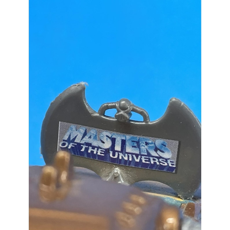 May include: A gray plastic accessory with the text "MASTERS OF THE UNIVERSE" in blue and white. The accessory has a bat-wing shape with a small handle at the top. The background is a bright blue.